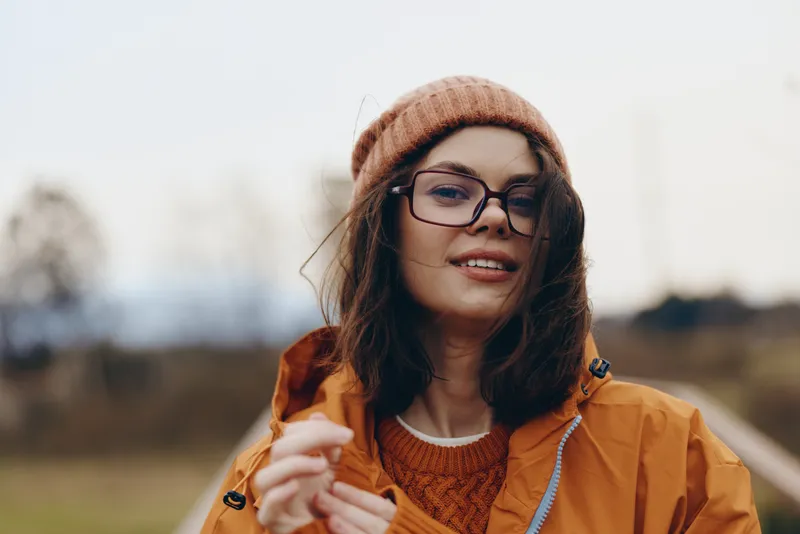 Woman lifestyle portrait outdoors, smiling brunette wearing orange jacket, knit hat and glasses, holding a coffee mug, soft sunlight, autumn nature background, relaxed casual mood