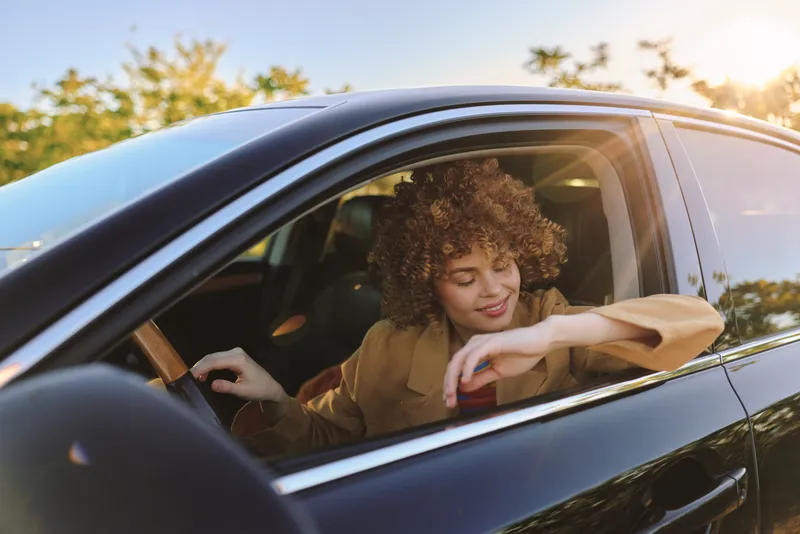 Cheerful woman inside a car, smiling with eyes closed, sunlit outdoor drive, relaxed posture, soft breeze, natural colors, lush greenery visible through window, warm afternoon, candid mood