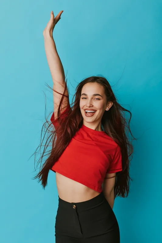 Young woman smiling with long hair wearing red crop top and black pants raising hand in excitement on blue background. Happiness, youth, casual style and positive energy concept.