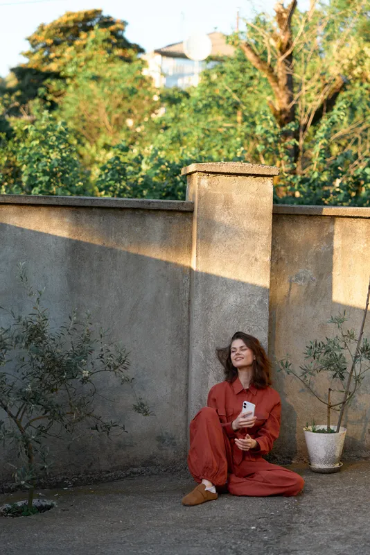 Cheerful woman outdoors in a red dress, relaxed sitting pose beside a courtyard wall, holding a coffee, sunlit mood, natural colors, tranquil garden backdrop, gentle breeze, candid charm