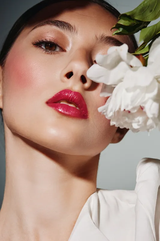 Close-up of a woman with glossy red lips and glowing skin holding white flowers near her face. Beauty and fashion concept with creative vibrant makeup and elegant attitude.