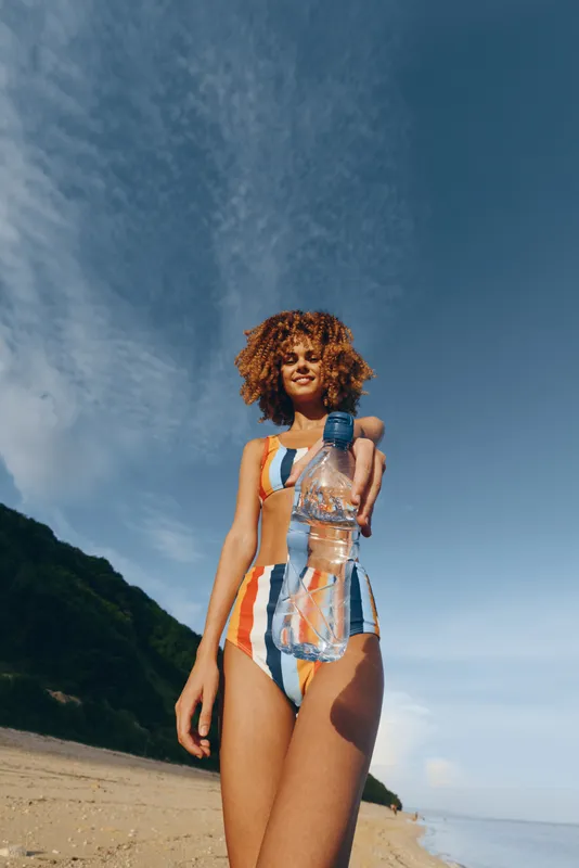 Woman standing on sandy beach, wearing colorful striped swimsuit, curly hair and sunlight, holding a bottle with water splash, ocean and clear sky behind, summer vacation lifestyle, outdoors portrait
