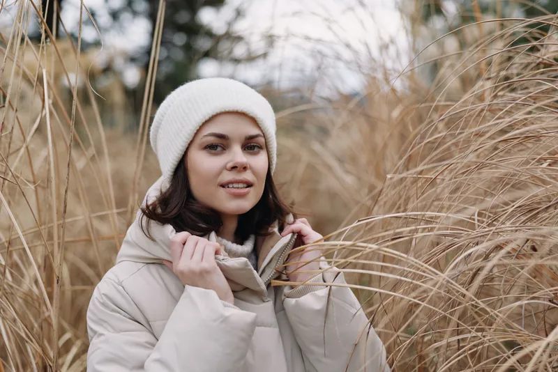 Woman in a light winter coat and white beanie standing among tall grass outdoors in soft daylight, showcasing cozy fashion, natural textures, and a calm seasonal mood with gentle surroundings.