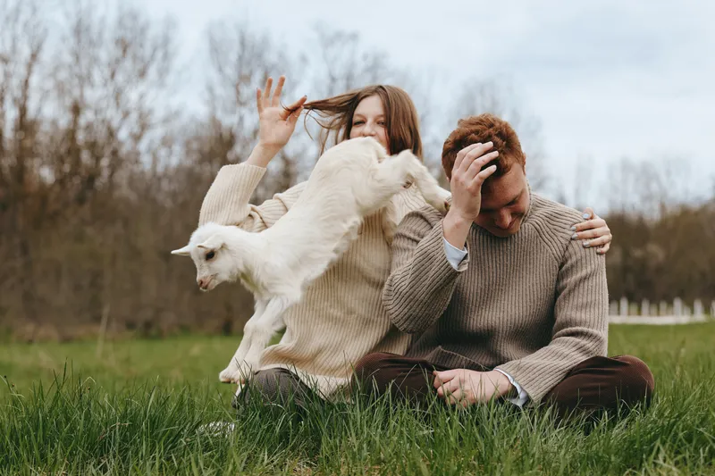 A couple sits on lush grass outdoors with a playful goat kid nearby, capturing a warm lifestyle moment of joy, bonding, and simple countryside happiness.