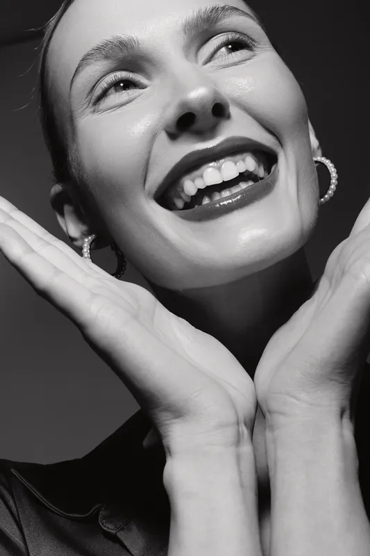 Close-up of a smiling white woman with fair skin and earrings posing with hands near face in studio lighting. Concept of happiness, beauty, and joyful female expression.