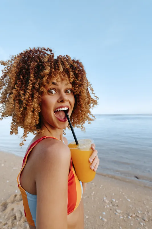 Cheerful woman curly hair beach ocean smiling holding drink with a straw, looking at camera, sunlit summer day, ocean horizon and waves behind, natural tones