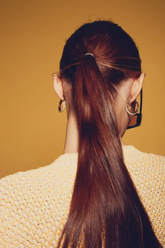 Back view of woman with long brown hair tied in ponytail wearing gold hoop earrings and yellow knit sweater against orange background. Concept of hairstyle and fashion accessories.