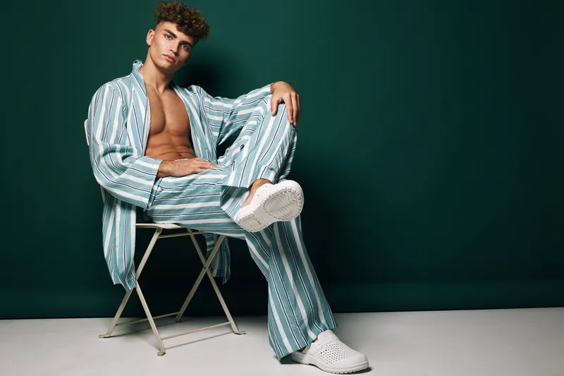 Young man sitting on chair wearing striped pajamas and white shoes against dark green background. Casual fashion, relaxed pose, male model with curly hair and bare chest in studio.
