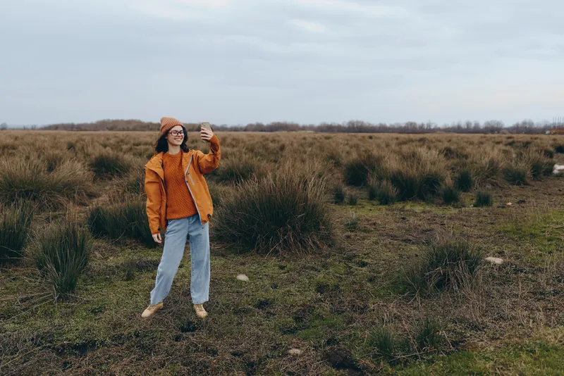 Woman outdoors in an autumn field, nature landscape, walking on a path, holding a smartphone up as if taking a photo, casual lifestyle portrait, soft natural daylight, calm mood