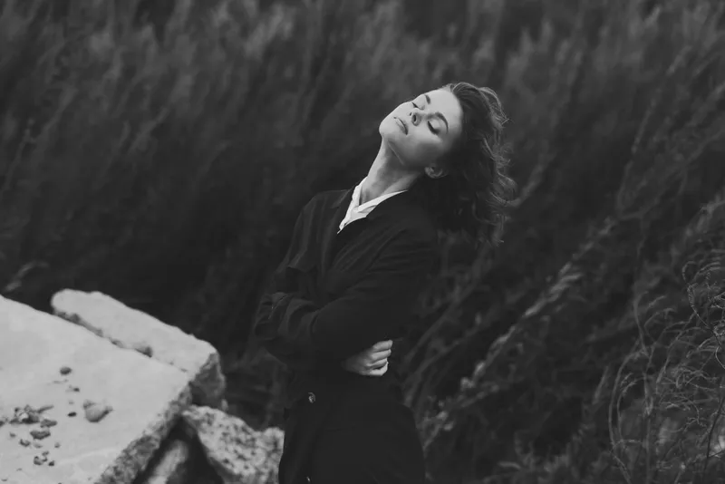 Woman in a dark coat outdoors, head tilted back, wind in her hair, serene pose among rocks, grass and a quiet landscape under muted light