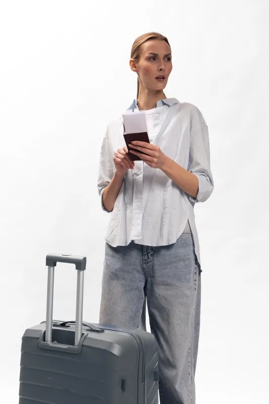 Young woman in casual clothes holds passport and ticket while standing near suitcase. Concept for travel, vacation planning, and airport waiting with focused expression on white background.