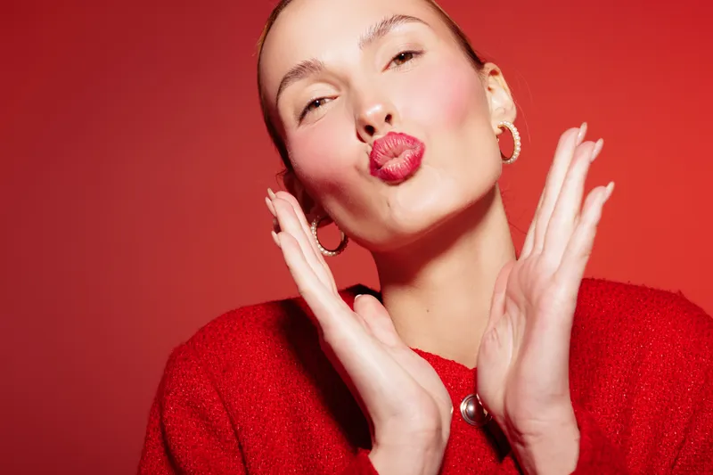 Young woman in red sweater sends a kiss with puckered lips and hands near face. Bright red background and hoop earrings create a vibrant, playful mood and fashion concept.