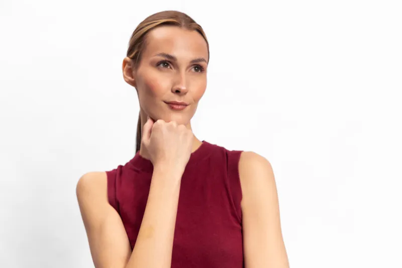 Thoughtful woman in sleeveless red top looks away with hand on chin against white background. Concept for contemplation, decision making, and calm confidence in portrait photography.