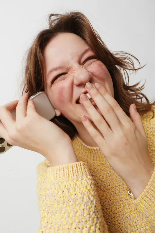Happy young woman in yellow sweater laughs joyfully while holding smartphone near ear. Close-up portrait on white background. Concept of communication, happiness, and casual lifestyle.