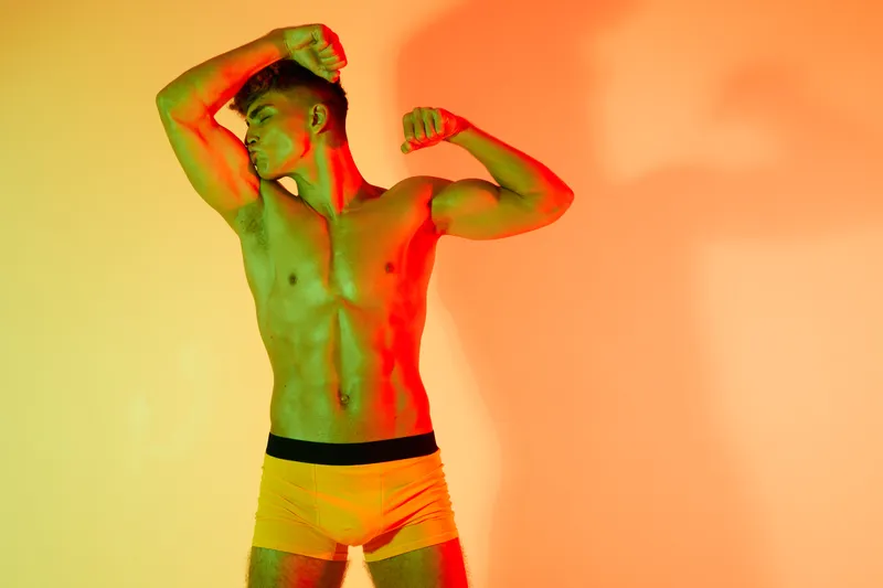 Muscular man posing in colorful lighting with flexed arms and bare torso wearing yellow underwear. Fitness, bodybuilder, strength and confidence in studio with artistic shadows and vibrant background.