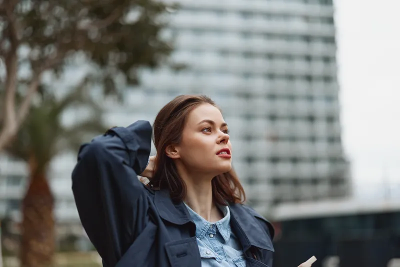 Young woman with brown hair stands outdoors in an urban setting, looking up with a thoughtful expression, wearing a navy trench over a denim shirt, surrounded by modern buildings.