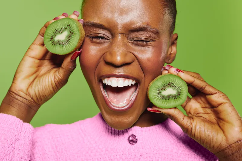 Joyful dark skinned African woman with closed eyes holding two halves of fresh kiwi fruit near face. Bright green background and pink sweater create vibrant cheerful mood and healthy lifestyle