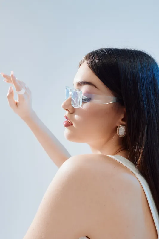 Young woman in transparent glasses poses with closed eyes and raised arm. Soft natural light highlights her beauty and fashion style in a minimalist setting.
