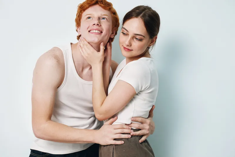 Young couple embracing and smiling with affection, woman gently holding man’s face, studio portrait with casual clothing and light background for relationship concept.