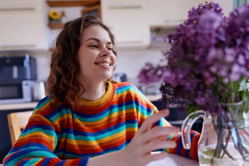 A smiling woman sits at a dining table in a colorful rainbow striped sweater, in a cozy home kitchen with flowers on the table, conveying cheerful, everyday lifestyle and warmth.