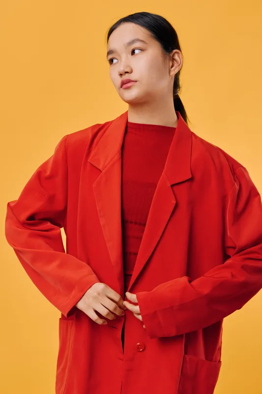 Young woman in red jacket and sweater standing against orange background looking sideways with serious expression and hands adjusting blazer for fashion portrait.