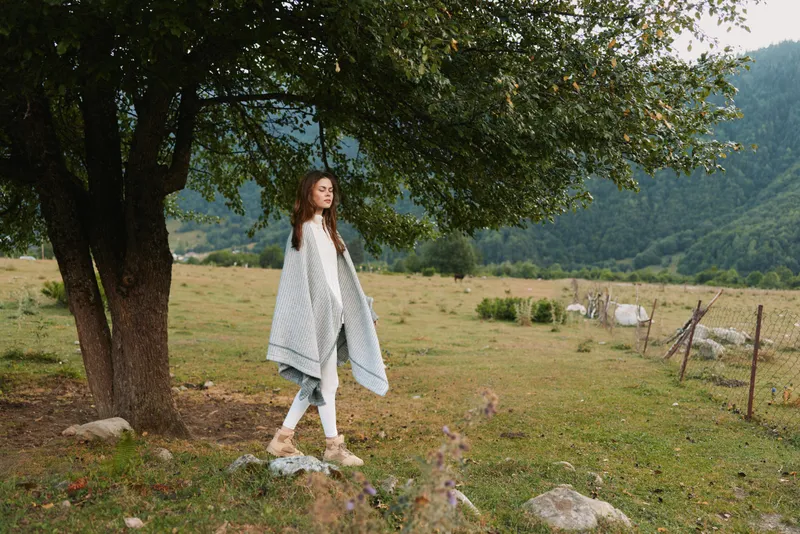 A woman walks through a grassy field under a large tree, wearing a light shawl. A peaceful countryside landscape with rolling hills and open skies conveys a calm lifestyle moment.