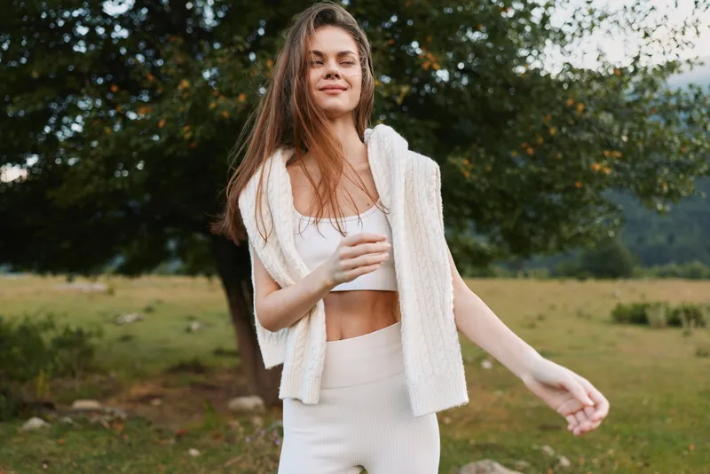 Young woman in a white two piece outfit stands in a meadow, smiling softly as a light cardigan drapes over her shoulders, creating a relaxed lifestyle moment