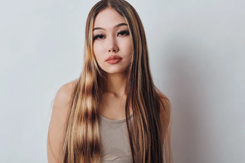 Young woman with long straight hair posing against white background with natural makeup and neutral expression wearing sleeveless top indoors.