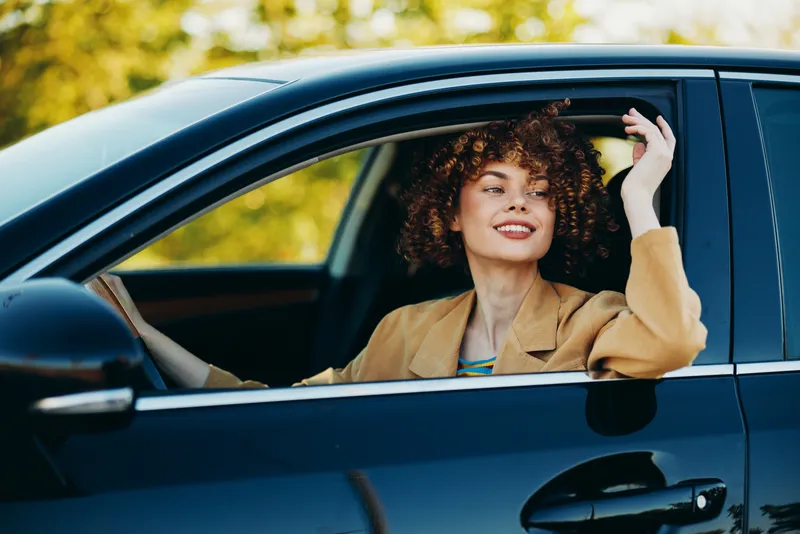 Smiling woman in a car with curly hair, retro film color enhances warm daylight mood. Outdoors in an urban setting, casual fashion and vintage vibes convey happiness and lasting peace.