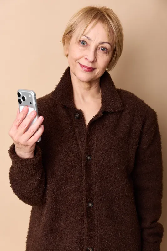 Woman holds smartphone for portrait, senior with smile in brown coat, looking at camera, beige studio background, technology communication, everyday life