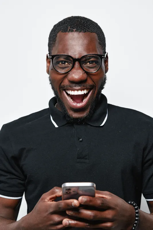 African American man with dark skin and glasses smiling widely while holding smartphone in black polo shirt on white background. Happy male using mobile phone for communication and technology.