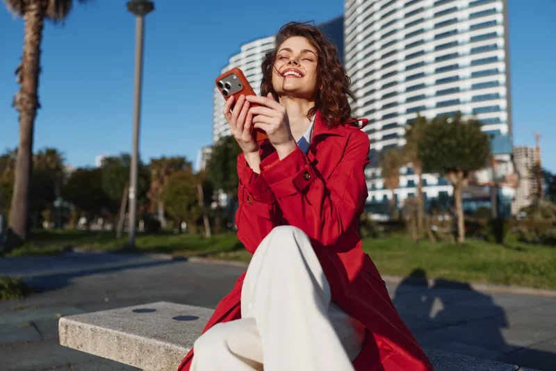 Smiling woman with curly hair wearing a red coat outdoors, shown with a smartphone. Urban park setting with modern buildings in the background. Casual fashion, happiness, and everyday tech moment.