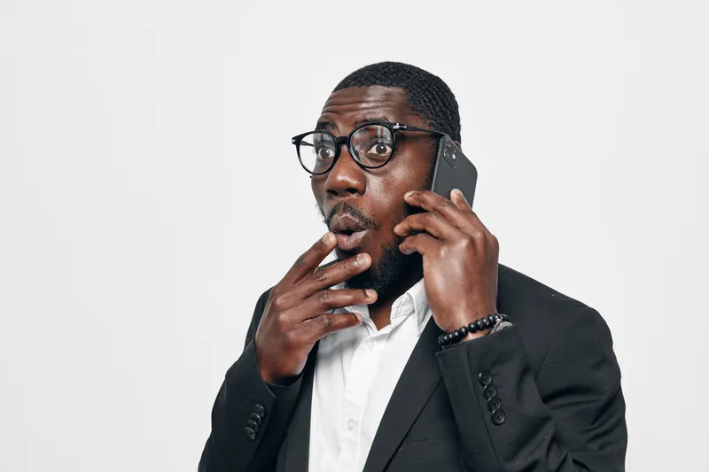 African American man with dark skin wearing glasses and black suit talking on smartphone with surprised expression isolated on white background. Business communication concept.