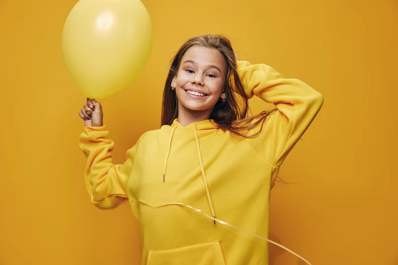 Girl smiling with balloon and yellow hoodie on yellow background for celebration, happiness and childhood joy with casual outfit and cheerful mood.