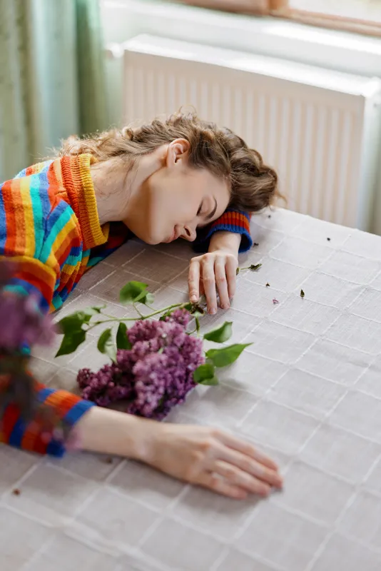 Cozy indoor moment with a woman resting her head on a table, colorful sweater, soft light, flowers nearby, calm intimate mood, home lifestyle scene, relaxed atmosphere