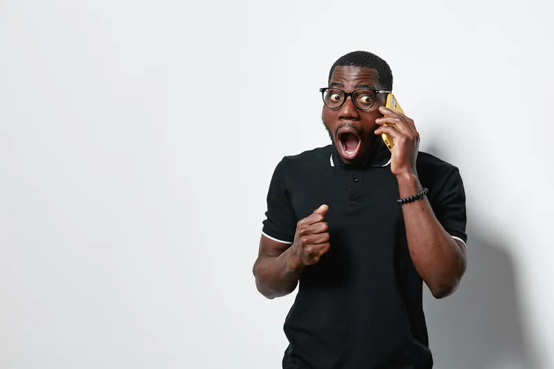 African American man with dark skin and glasses talking on smartphone with surprised expression in casual black shirt against white background. Communication, emotion and technology.