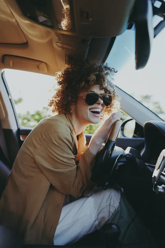 A woman driver with curly hair and sunglasses smiles in a sunlit car, capturing a carefree lifestyle moment with retro film color and candid warmth, concept for travel and spontaneity.