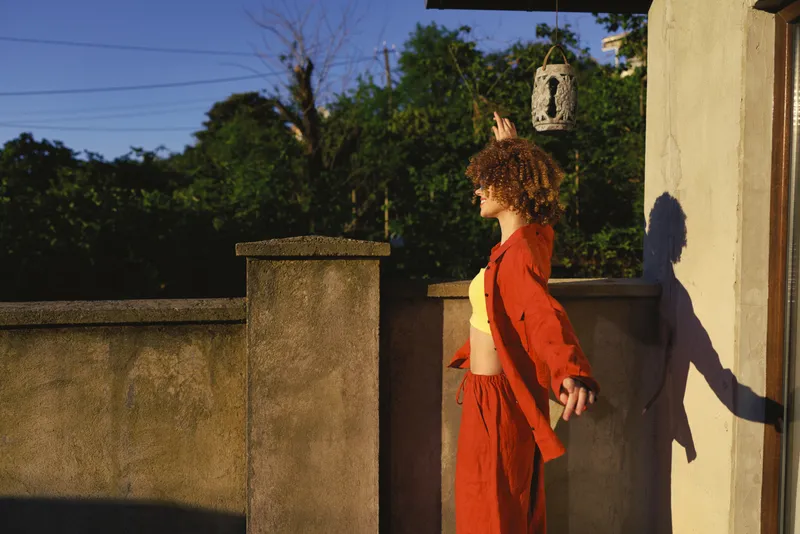 Young woman with curly hair in a bright red outfit stands on a sunlit terrace, stretching her arms as a warm shadow stretches along the concrete wall, exuding confidence
