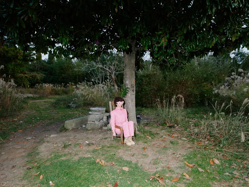 Child girl sits on a small chair beneath a large tree in a quiet park, wearing a pink dress, surrounded by grass, leaves and natural greenery in daylight during a calm afternoon.