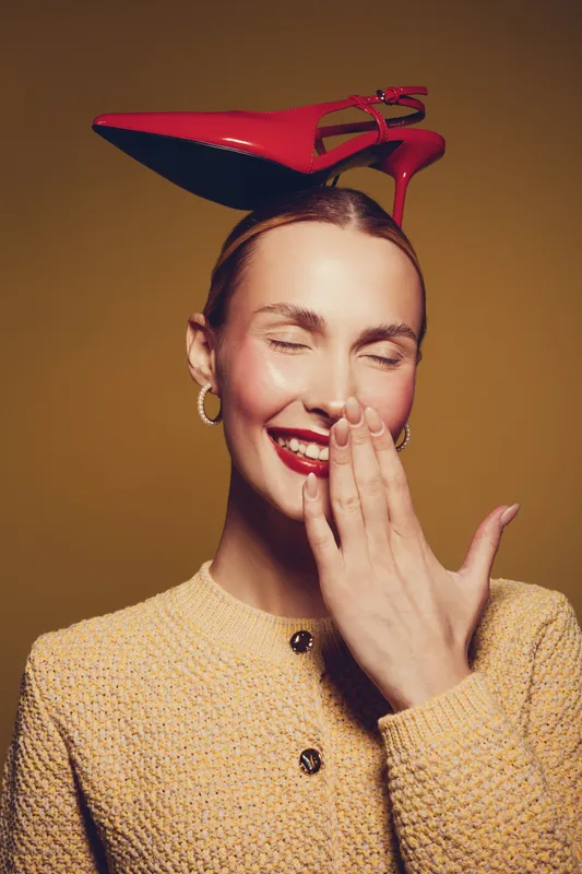 Smiling woman with red high heel shoe on head covers mouth with hand, wearing yellow textured jacket. Studio portrait on brown background. Concept of fashion, humor, and playful style.