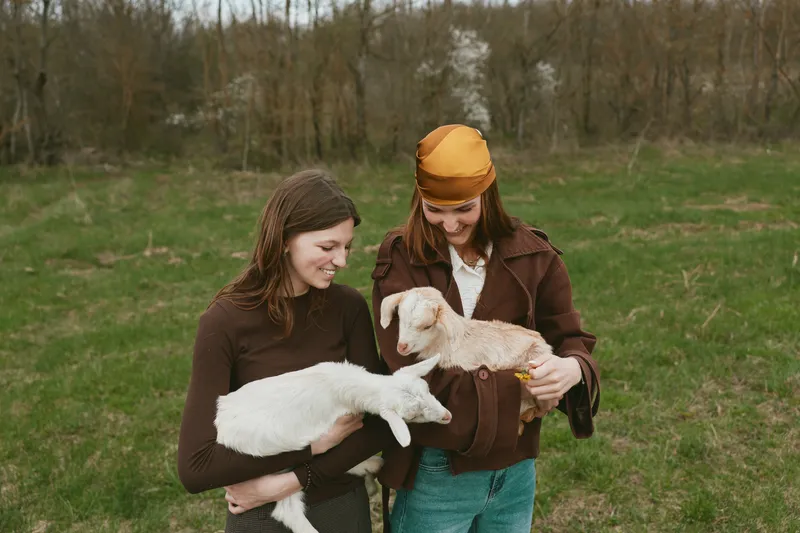 Two women outdoors share a gentle moment with a young goat kid, smiling as they cradle it. A lifestyle scene highlighting animal companionship, care, and countryside joy.
