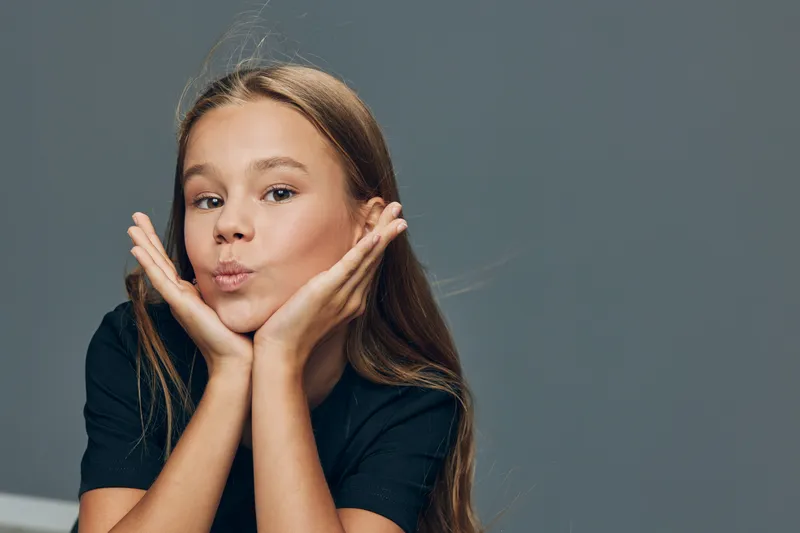 Girl with long hair and black shirt posing with hands face closeup, smiling and playful expression with grey background in studio portrait photography.
