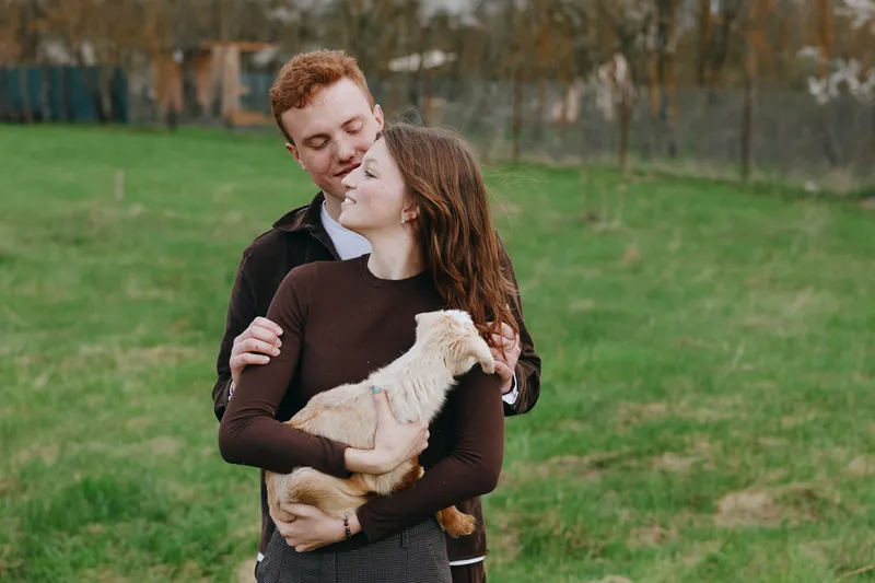 Young couple embraces outdoors as woman holds a small goat in her arms while they enjoy a peaceful moment together in a green grassy field during autumn.