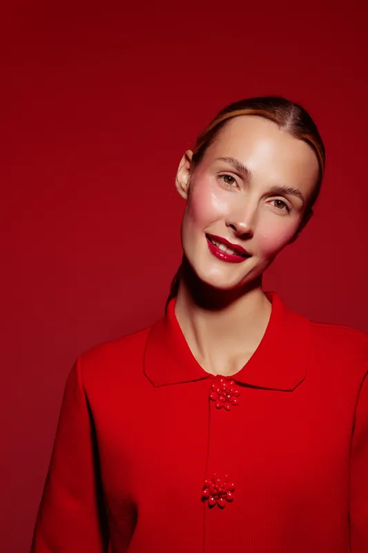 Young woman with red lipstick and red sweater smiles gently against red background. Studio portrait with soft lighting highlights natural skin texture and warm expression.