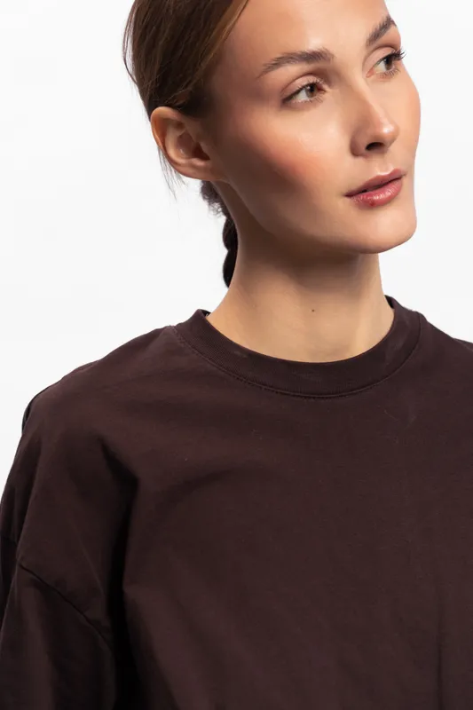 Young woman in dark brown shirt looks sideways with calm expression on white background. Minimalist portrait with natural light and soft shadows. Concept of casual style and simplicity.