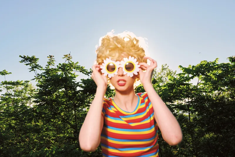 Woman with blonde curls wearing a striped shirt, holding playful flower sunglasses outdoors. Retro film color, lifestyle vibe, sunny mood and cheerful energy, natural light enhances textures.