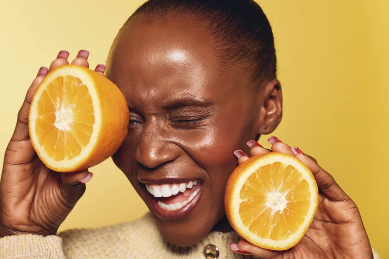 Happy African woman with dark skin holding two orange halves near her face, smiling with closed eyes on yellow background. Concept of fruit, health, and natural beauty.