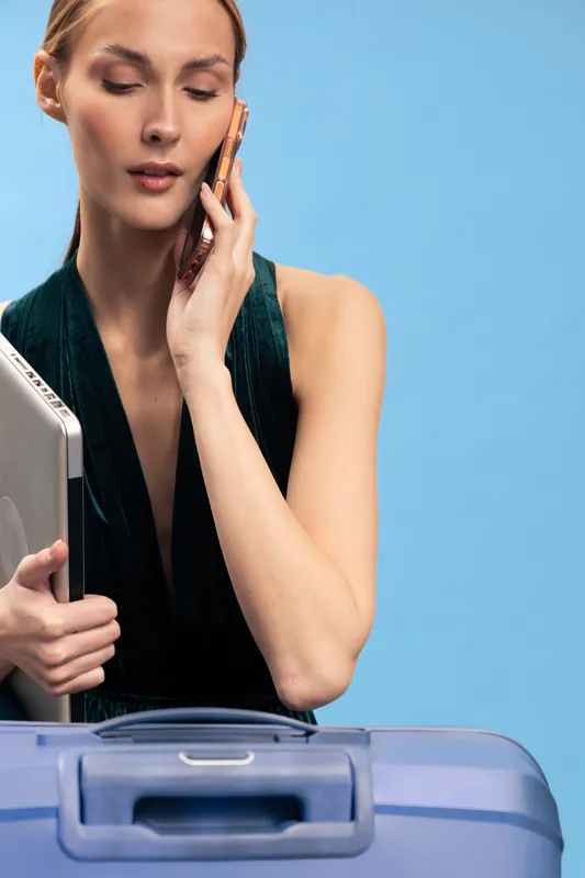Young woman holds laptop and talks on smartphone with serious expression. Blue background and suitcase in foreground. Concept of travel, work, business, and communication.