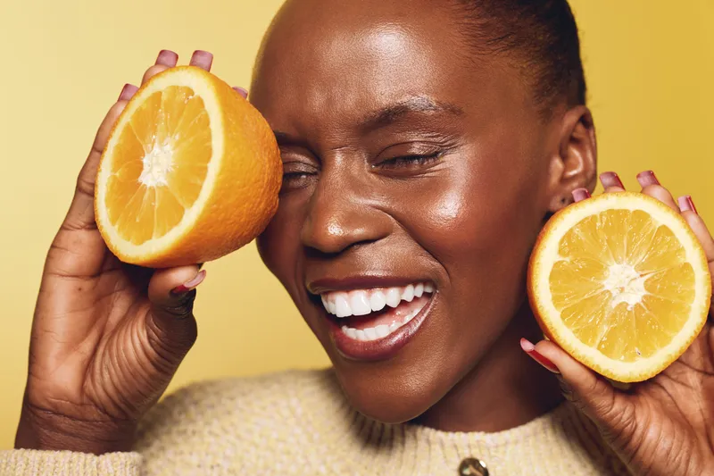Happy dark skinned African woman with closed eyes smiling and holding two halves of fresh orange fruit near face. Concept of healthy lifestyle, vitamin, wellness, and natural beauty.
