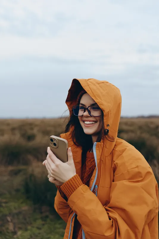 Smiling woman outdoors, hooded orange jacket, sunglasses on, holding smartphone in her hands, rural field background, lifestyle travel feeling, nature spring portrait in soft daylight, relaxed smile